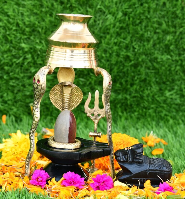 A stone Shiva Lingam sits on a black base under a brass cobra-shaped stand, accompanied by a small Nandi statue and marigold flowers on green grass.