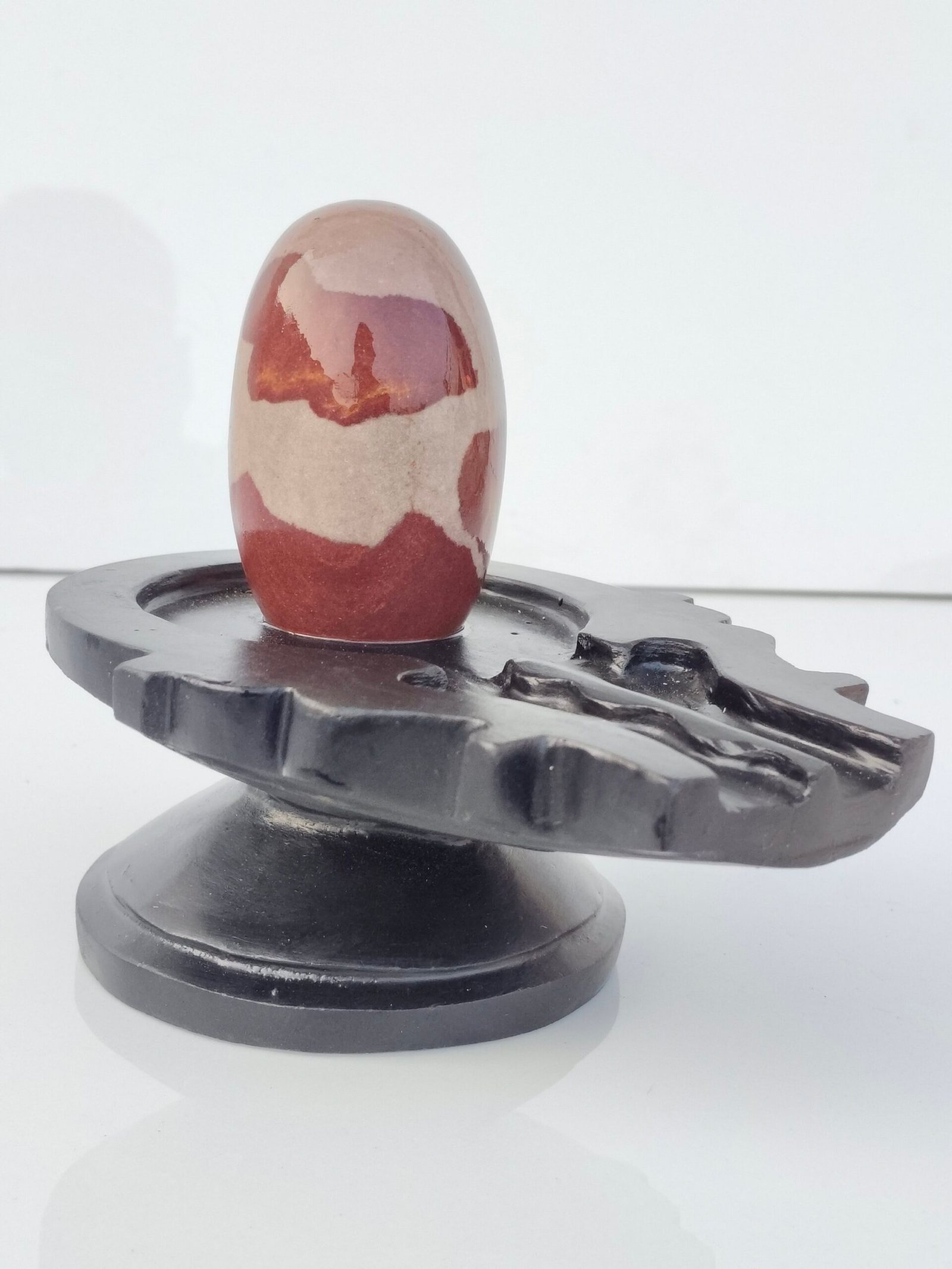 A smooth, oval-shaped Narmadeshwar Shivling with reddish-brown and tan patterns, resting on a black stone jaladhari base.