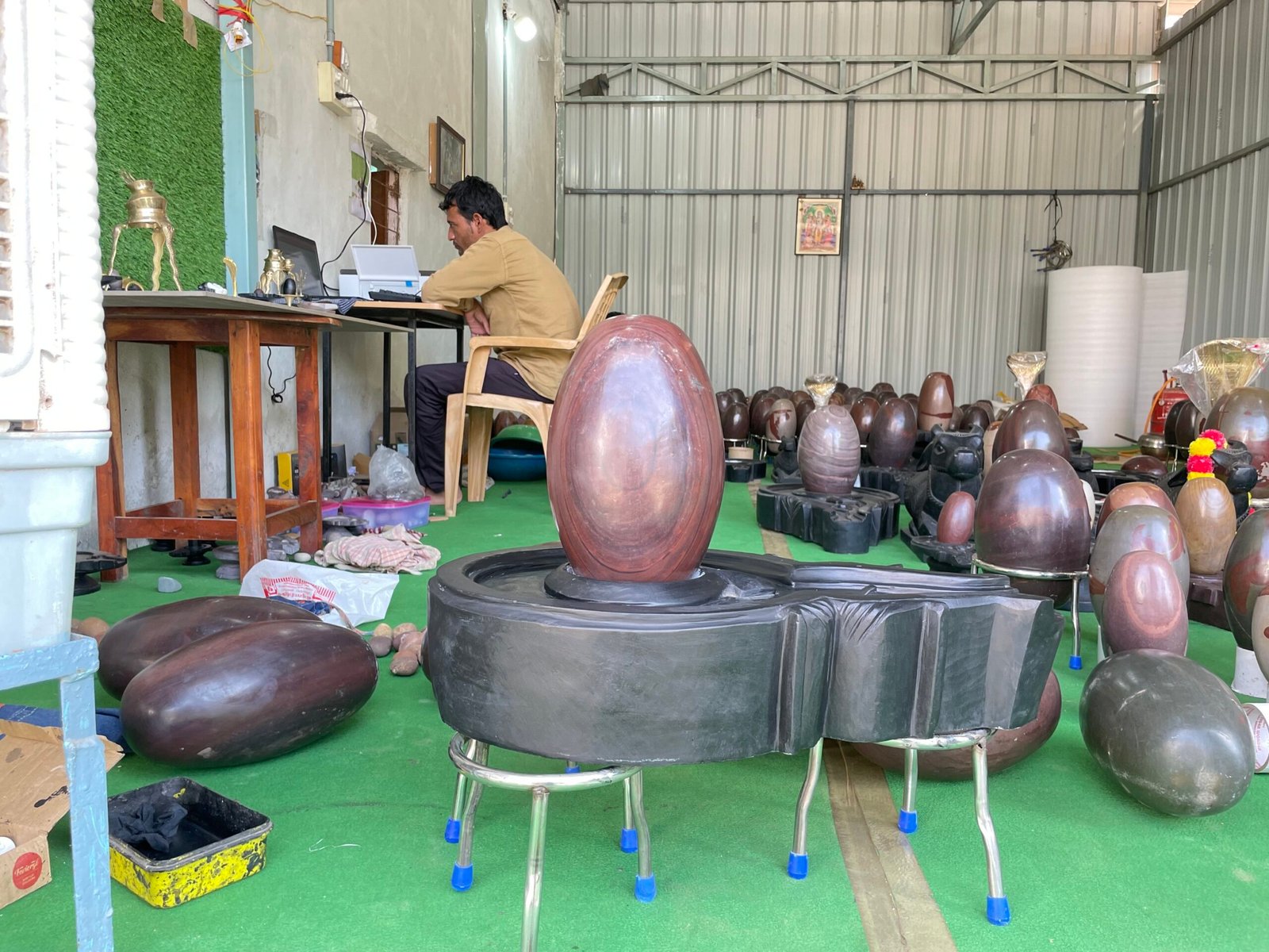 A craftsman works at a desk in a workshop filled with various polished stone lingam sculptures.