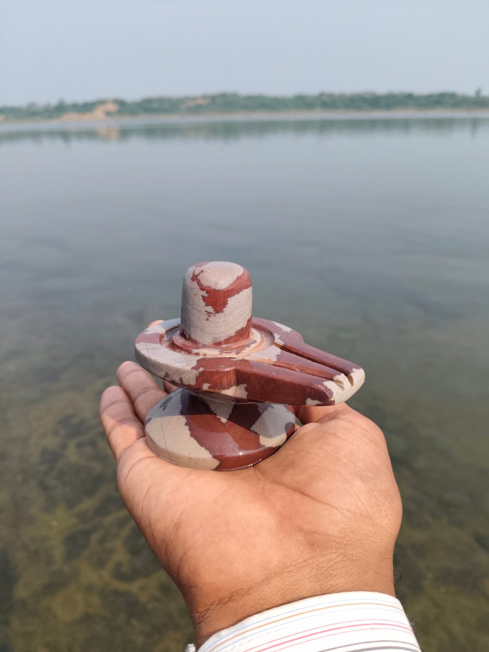 A person holds a small, polished stone Shiva Lingam with red and beige patterns against a blurred background of a calm river.