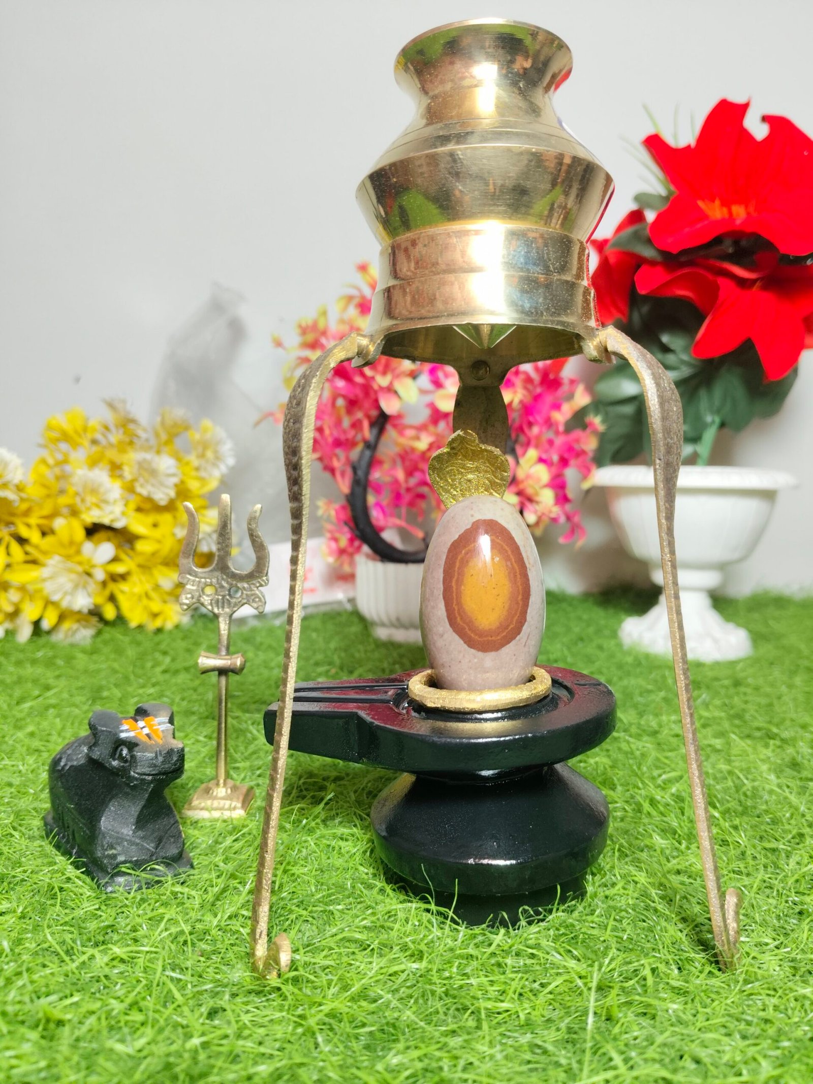 A stone Shiva Lingam sits on a black base under a brass canopy, accompanied by a small Nandi statue and a miniature trishula on a green surface.