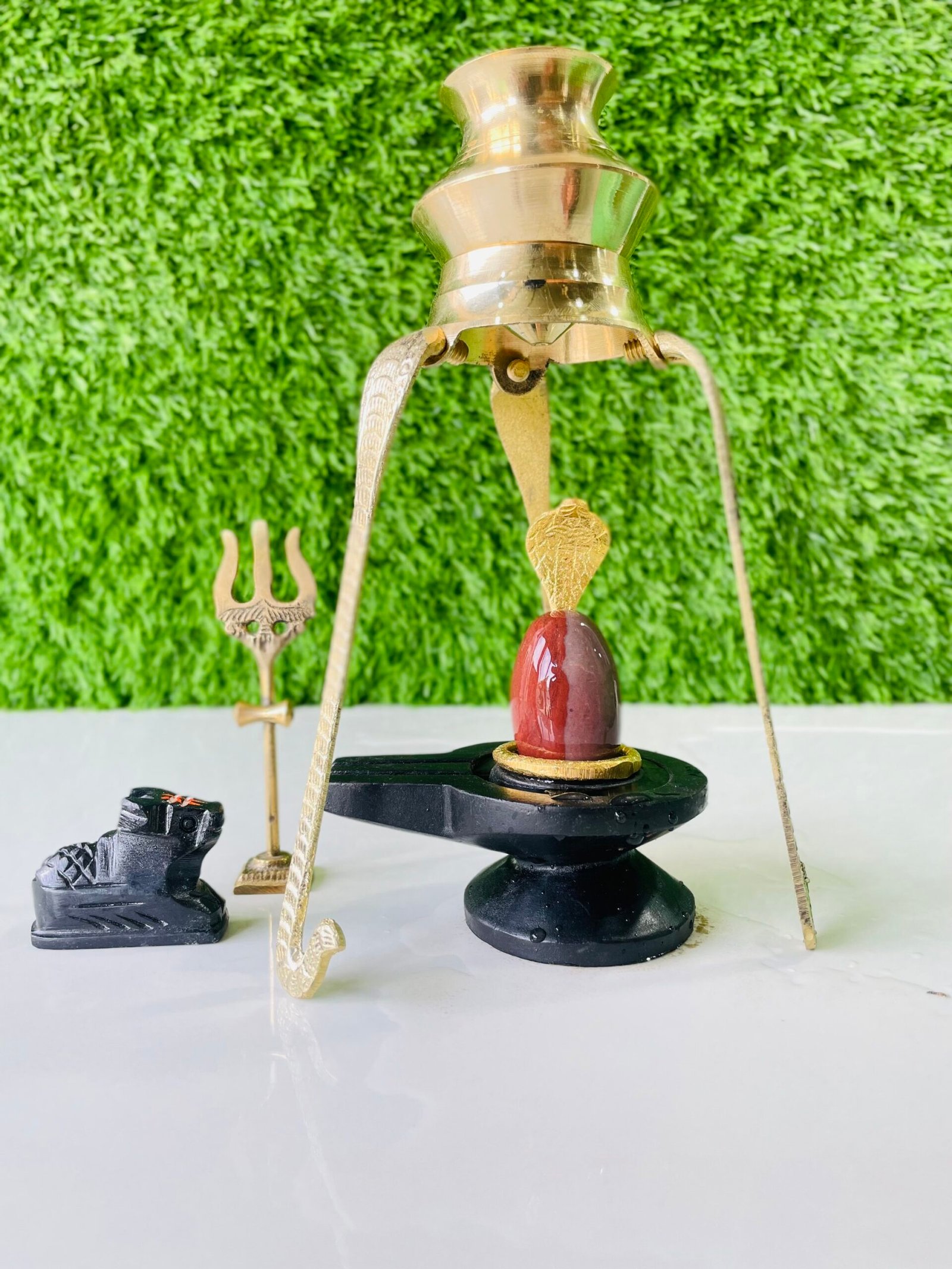 A Narmadeshwar Shivling with a brass abhishekam stand, a small Nandi statue, and a trishul arranged on a white surface against a green background.