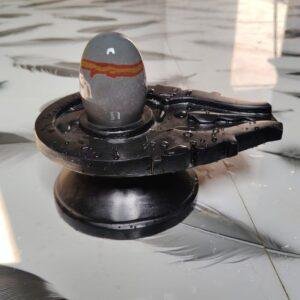 A smooth, grey stone Shivling with a horizontal orange stripe placed on a black, water-dampened Jaladhari base.