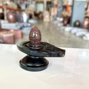 A polished, reddish-brown stone Shiva Lingam sits centered on a black, carved Yoni base against a blurred background.