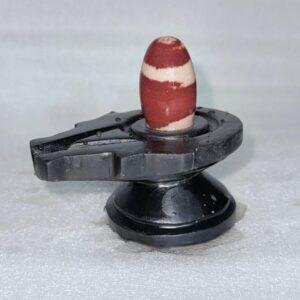 A smooth, oval-shaped red and white banded jasper stone resting upright in the center of a black, carved yoni pedestal.