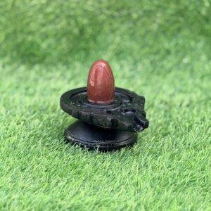A small, polished reddish-brown stone Shivling placed on a black, circular Damru-shaped Jaladhari base, set against a green grass background.