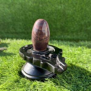 A polished, brown-striped stone Shivling placed on a black, circular jaladhari base, set against a green artificial grass background.
