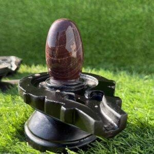 A smooth, brown, oval-shaped stone Shivling placed on a black, carved stone jaladhari base, set against a background of green artificial grass.