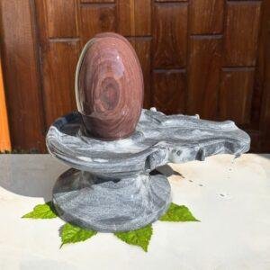 A smooth, brown, oval-shaped Shivling placed on a grey, carved stone Jaladhari base with water dripping from the spout.