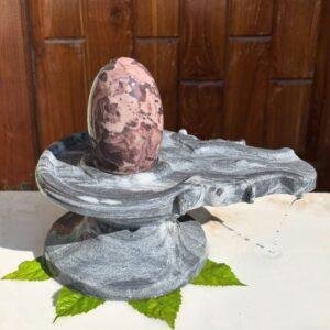A polished, mottled pink and brown stone Shivling resting on a grey stone Jaladhari base with water dripping from the spout.