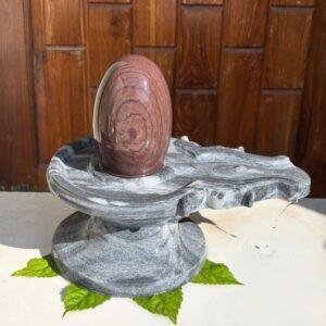 A smooth, oval-shaped brown stone Shivling resting on a grey marble Jaladhari base, placed on a white surface with green leaves.