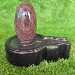 A polished, oval-shaped brown and reddish stone Shivling resting on a dark, circular stone jaladhari base, set against a green artificial grass background.
